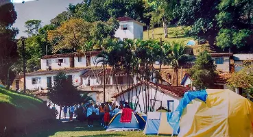 Foto colorida de um gramado no Morrinho com diversas barracas de camping armadas em frente a casas e ranchos construídos na encosta da colina.