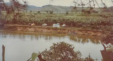 Foto colorida de 1982 do Rio Juquiá. Carros e uma Kombi estão estacionados na margem, próximos a um barranco. No rio, um pequeno grupo de pessoas atravessa a água em um barco.