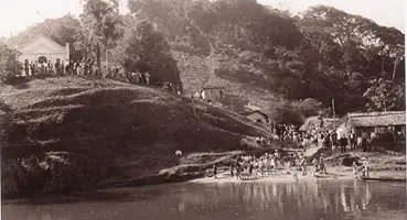 Foto histórica em preto e branco do Morrinho, com o templo no topo de uma colina íngreme e uma grande multidão de pessoas reunida na margem do Rio Juquiá, o ponto de desembarque